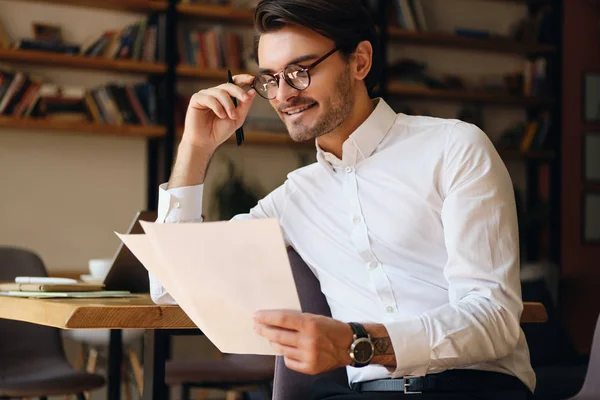 Joven hombre de negocios sonriente en gafas y camisa blanca felizmente trabajando con papeles en la oficina moderna — Foto de Stock