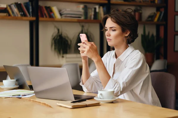 Молодая задумчивая женщина сидит за столом с ноутбуком и чашкой кофе задумчиво используя мобильный телефон в современном офисе — стоковое фото