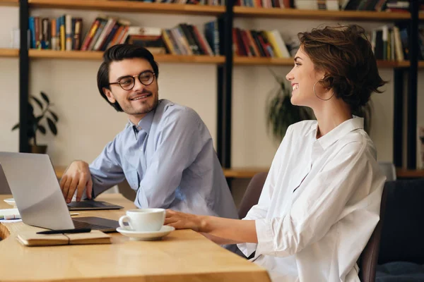 Νέοι χαρούμενοι επιχειρηματικοί συνάδελφοι που κάθονται στο τραπέζι με το laptop να μιλούν ευχαρίστως στη δουλειά στο σύγχρονο γραφείο — Φωτογραφία Αρχείου