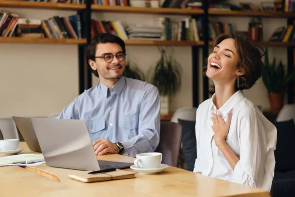 Modern ofiste iş yerinde sevinçle konuşan dizüstü bilgisayar ile masada oturan genç gülümseyen iş arkadaşları — Stok fotoğraf