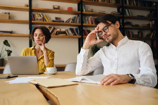 Ung trött man i glasögon sitter vid bordet lutande huvudet på hand eftertänksamt arbetar på kontoret med kollega på bakgrunden — Stockfoto