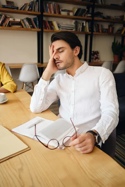 Junger müder Mann sitzt am Tisch mit Notizblock, den Kopf auf die Hand gestützt und schläft bei der Arbeit im modernen Büro — Stockfoto