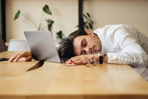 Junger müder Mann mit Laptop schläft auf Schreibtisch bei der Arbeit im modernen Büro — Stockfoto