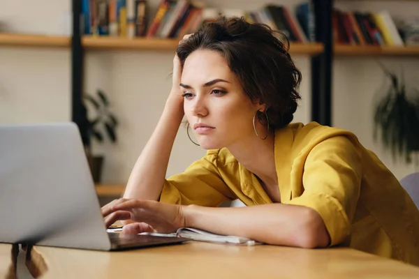 Joven mujer molesta sentada en el escritorio trabajando cansadamente en un nuevo proyecto con computadora portátil en la oficina moderna —  Fotos de Stock