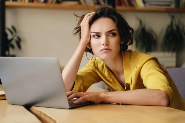 Porträtt av unga seriösa affärskvinna sitter vid skrivbordet tiredly ser åt sidan arbeta på laptop i moderna kontor — Stockfoto