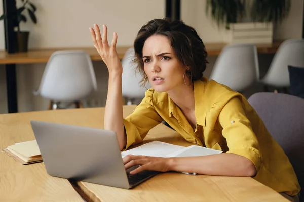 Young disappointed businesswoman sitting at the desk with laptop indignantly looking aside at work in modern office