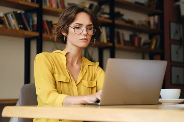 Jonge serieuze zakenvrouw in brillen zittend aan het Bureau doordacht werken aan nieuw project met laptop in modern kantoor Stockfoto