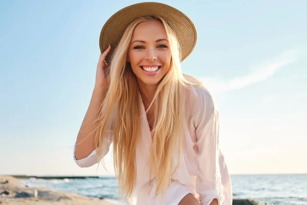 Young attractive smiling blond woman in white shirt and hat joyfully looking in camera with sea on background — Stock Photo, Image