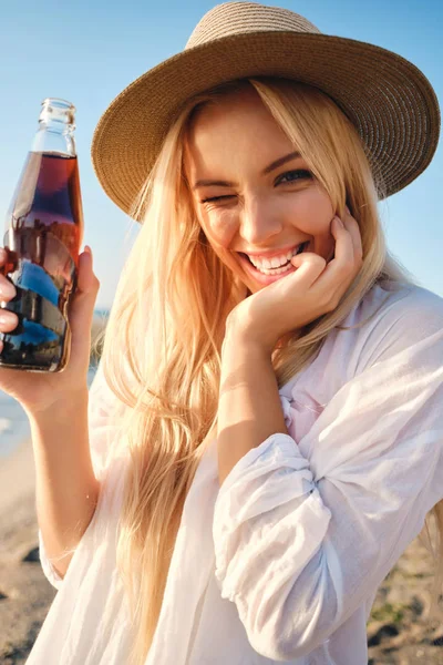 Молода радісна блондинка в капелюсі тримає пляшку содової води, щасливо підморгнувши, дивлячись в камеру біля моря — стокове фото