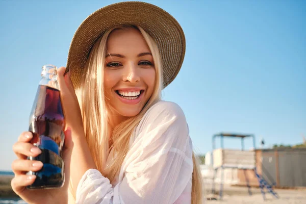 Young pretty smiling blond woman in hat holding glass bottle with soda water happily looking in camera on beach — Stock Photo, Image