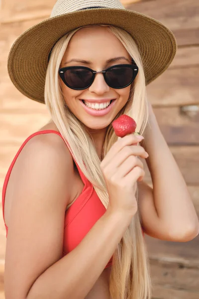 Junge hübsche, lächelnde blonde Frau im Badeanzug mit Sonnenbrille und Hut, die freudig Erdbeere am Strand hält — Stockfoto