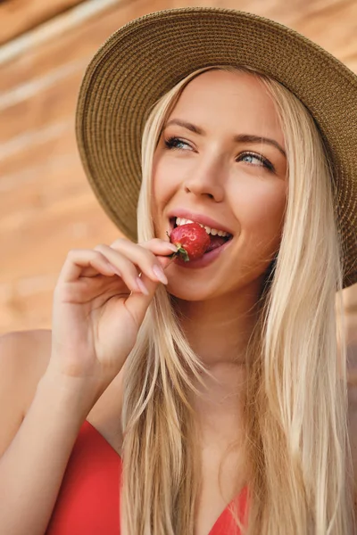 Potret wanita muda berambut pirang yang menarik dengan pakaian renang dan topi dengan mimpi melihat ke samping makan strawberry di pantai — Stok Foto