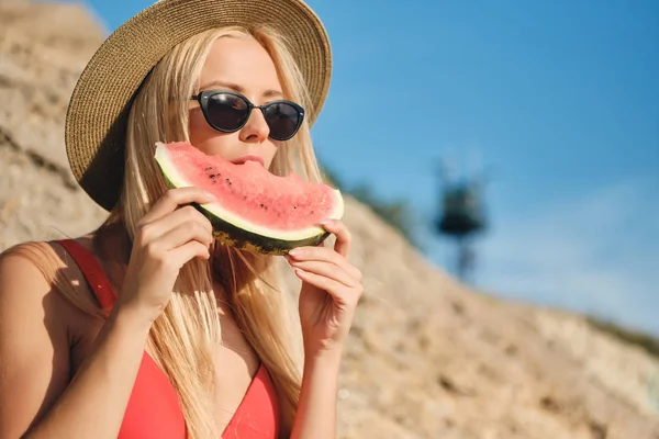 年轻的迷人的金发女郎穿着泳衣，戴着太阳镜和帽子，在海滩上梦寐以过地吃西瓜片的肖像 — 图库照片