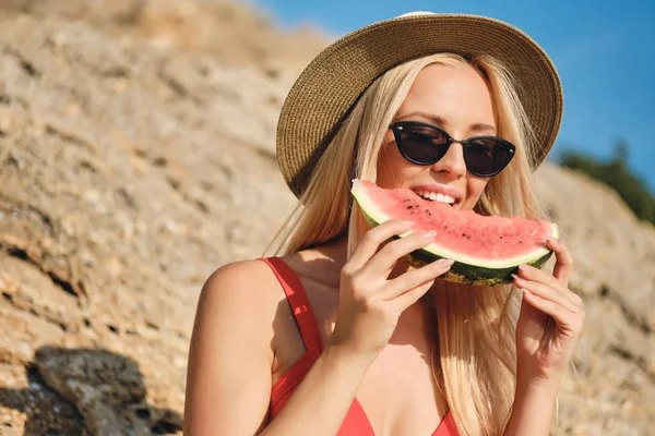 Portrait of young pretty blond woman in swimsuit wearing sunglasses and hat happily eating watermelon on beach — Stock Photo, Image