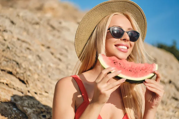 Portrait of young beautiful smiling blond woman in swimsuit wearing sunglasses and hat happily sitting with slice of watermelon on beach — Stock Photo, Image