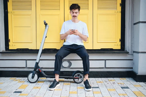 Ung stilig mustasch man sitter med e-Scooter eftertänksamt använder mobiltelefon med gul bakgrund på City Street — Stockfoto
