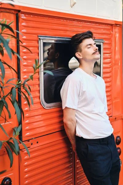 Side view of young handsome mustache man in withe T-shirt dreamily standing with eyes close near red van on city street — Stock Photo, Image