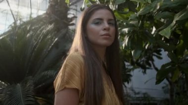 Tropik serada kameraya mutlu bir şekilde bakan çiçekli elbisesi olan güzel kahverengi saçlı kızın fotoğrafını takip etmek