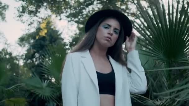 帽子とジャケットで魅力的なファッションの女の子のショットを追跡し 古い庭で大きな熱帯の葉とカメラを思慮深く見て — ストック動画