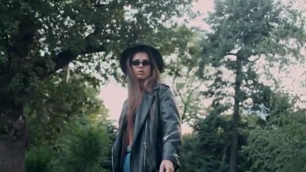 跟踪周围拍摄的有吸引力的自信的女孩在皮夹克和太阳镜周到脱下帽子走在老花园 — 图库视频影像