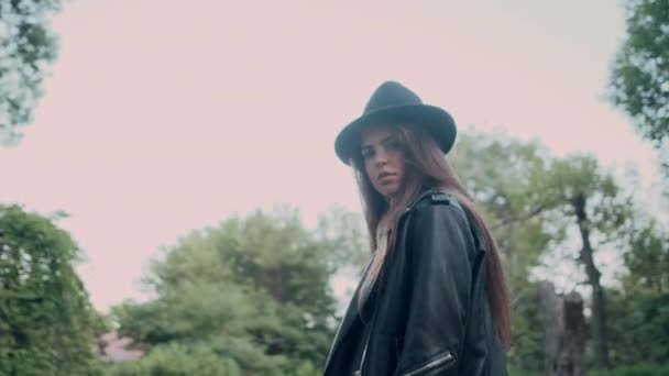古い庭を歩くカメラで自信を持って見ている革のジャケットと帽子で魅力的な女の子のショットの周りを追跡 — ストック動画