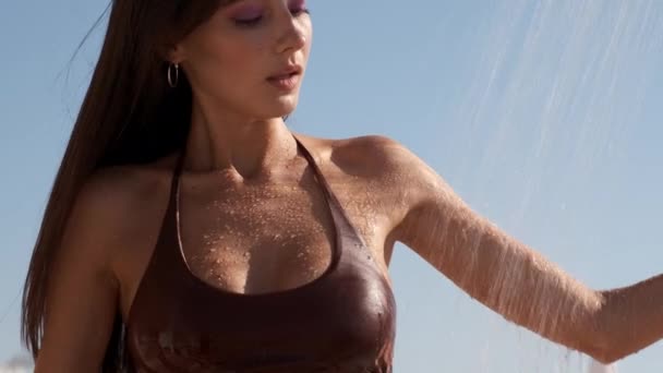 ビーチでシャワーの下に立って濡れた水着で魅力的なスリムなブルネットの女の子のクローズアップトラッキングショット — ストック動画
