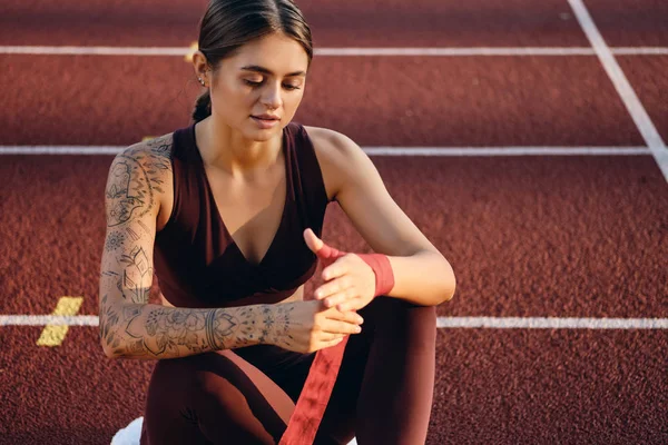 Beautiful sporty girl with tattooed hand in sportswear dreamily wrapping red boxing bandage on hand on stadium — Stock Photo, Image