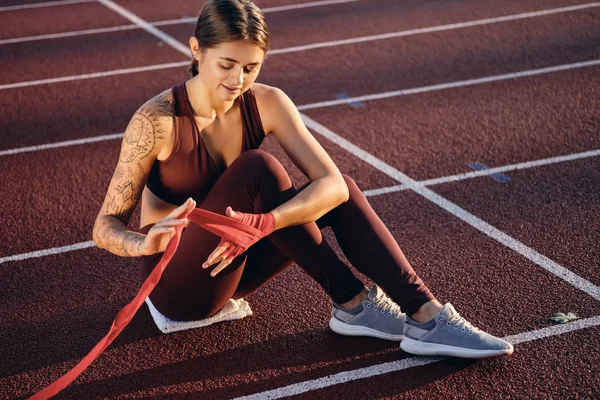 Attractive athlete girl with tattooed hand in sportswear wrapping red boxing bandage on hand on stadium — Stock Photo, Image