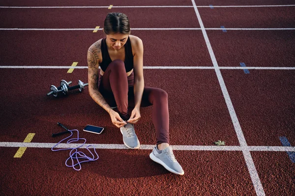 Attraktives Sportlermädchen mit tätowierter Hand in Sportbekleidung, das Turnschuhschnürsenkel bindet und sich auf das Training im Stadion vorbereitet — Stockfoto