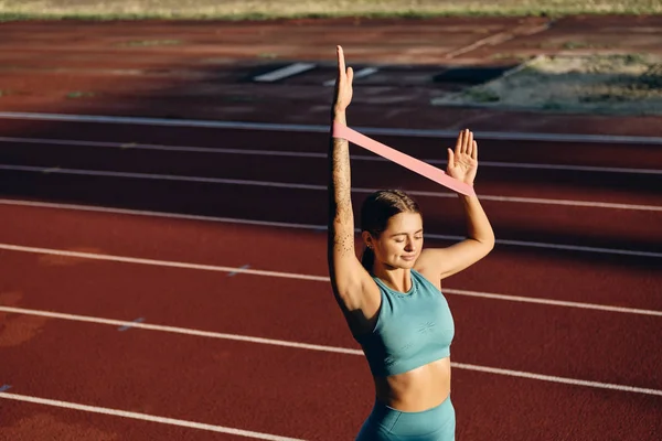 Bella ragazza atleta sorridente in elegante abbigliamento sportivo tenendo le mani con elastico durante l'allenamento sullo stadio della città — Foto Stock