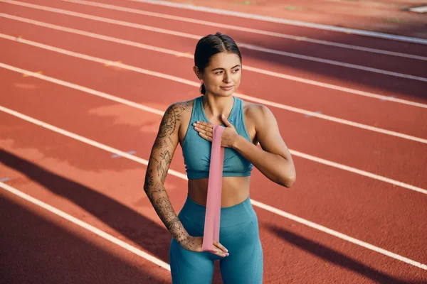 Csinos lány a stílusos sportruházat álmodosan stretching kezet gumiszalag edzés közben a városi stadionban — Stock Fotó