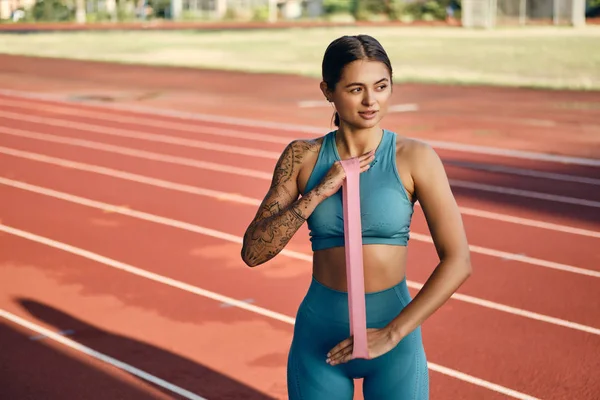 Attractive sporty girl in stylish sportswear stretching hands with rubber band during training on city stadium — Stock Photo, Image