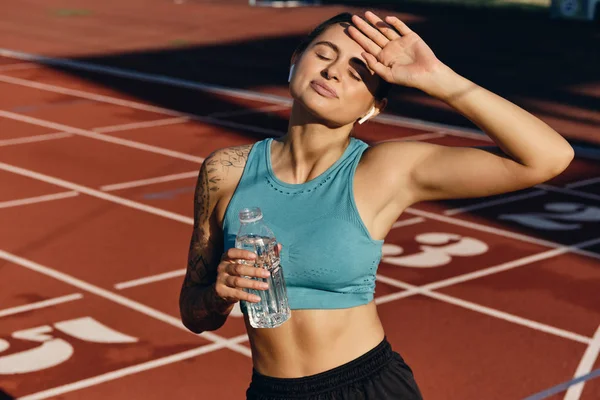 Tired sporty girl in top with bottle of water covering eyes with hand from sun after workout on city stadium — Stock Photo, Image