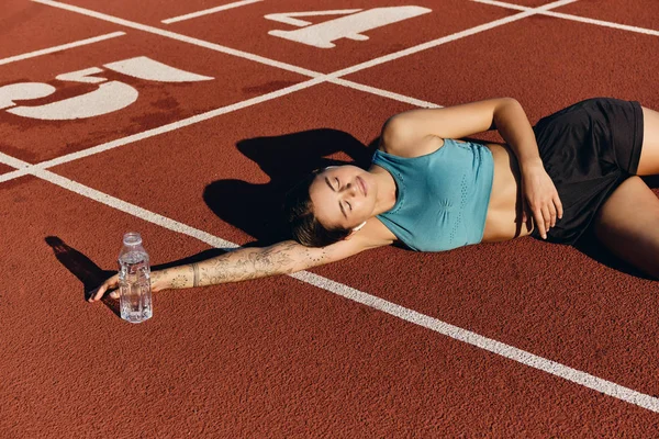 Mooie atleet meisje in sportkleding dromerige liggend op runner track met fles na training op stadion — Stockfoto