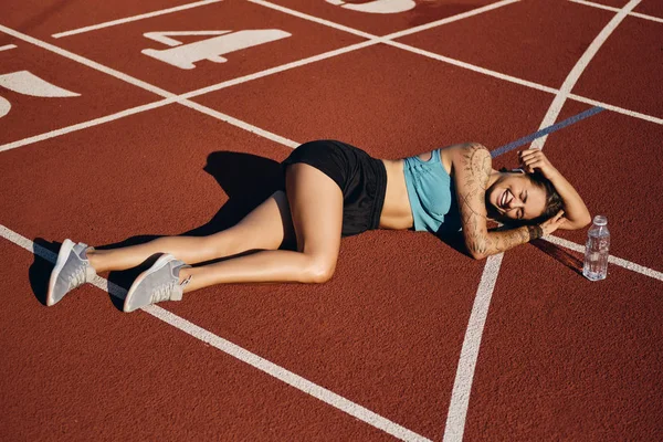 Весела спортивна дівчина в спортивному одязі радісно лежить на бігунній доріжці з пляшкою після тренування на стадіоні — стокове фото