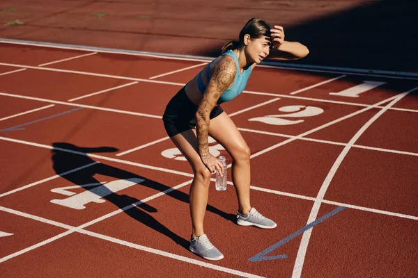 Young athlete woman in sportswear with bottle of water tiredly leaning on knees after running on city stadium — Stock Photo, Image