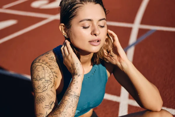 Cerca atractiva chica atleta mojada con la mano tatuada en ropa deportiva descansando soñadoramente después del entrenamiento en el estadio de la ciudad —  Fotos de Stock