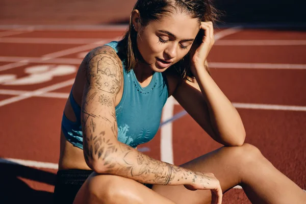 Σκεπτική υγρή αθλήτρια κορίτσι με τατουάζ χέρι σε αθλητικά είδη που ξεκουράζονται μετά από τρέξιμο στο στάδιο της πόλης — Φωτογραφία Αρχείου