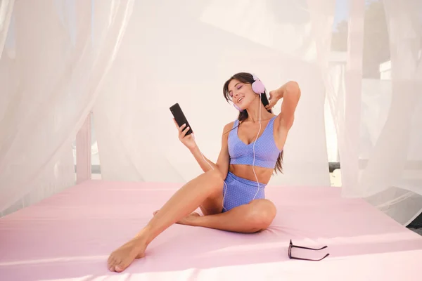 Atractiva chica morena sonriente en traje de baño lindo escuchando música en los auriculares en la cama de playa con cortinas transparentes —  Fotos de Stock