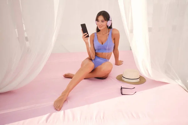 Mooie brunette meisje in schattig badpak en koptelefoon gelukkig met behulp van mobiele telefoon op het strand bed met gordijnen rond — Stockfoto