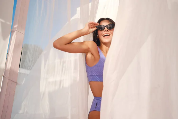 Vrolijk brunette meisje in schattig badpak en zonnebril vreugdevol op zoek in de camera met gordijnen rond aan zee — Stockfoto