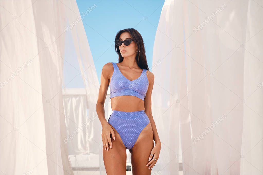 Gorgeous brunette girl in cute swimsuit and sunglasses thoughtfully standing around curtains fluttering in the wind on seaside