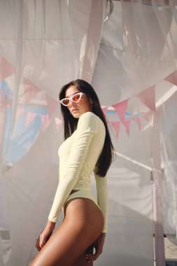 Mayo ve renkli güneş gözlüğü ciddi şık esmer kız düşünceli etrafında perdeler ile plaj yatağında kamera bakıyor