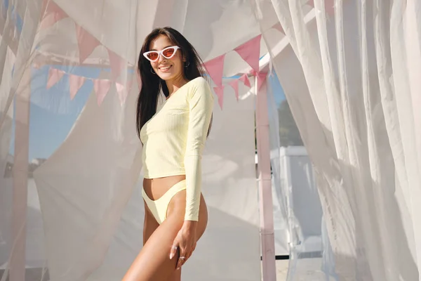 Cheerful stylish brunette girl in swimwear and colorful sunglasses happily looking in camera on beach bed with curtains around — Stock Photo, Image