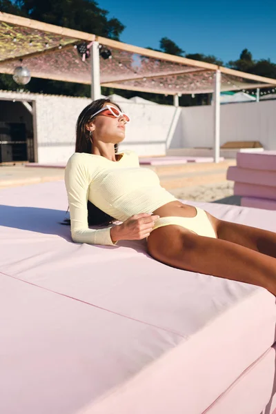 Beautiful brunette girl in swimwear and colorful sunglasses sensually sunbathing on pink beach bed — Stock Photo, Image