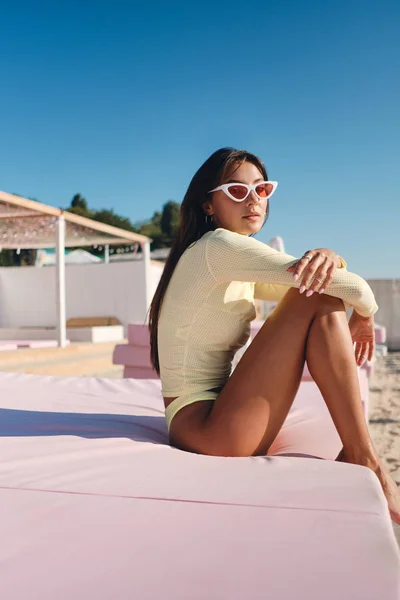 Beautiful stylish brunette girl in swimwear and colorful sunglasses dreamily looking in camera on pink beach bed — Stock Photo, Image