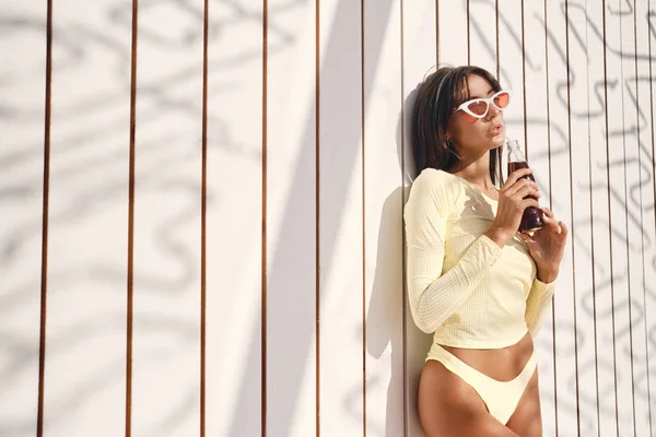 Mooie brunette meisje in stijlvolle badmode en kleurrijke zonnebril sensueel drinken soda over hout achtergrond op strand — Stockfoto
