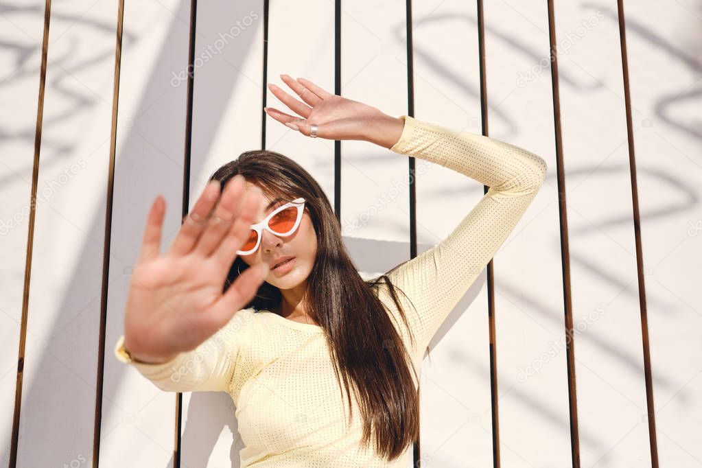 Gorgeous stylish brunette girl in swimwear and colorful sunglasses thoughtfully covering camera with hand posing over wood background on beach