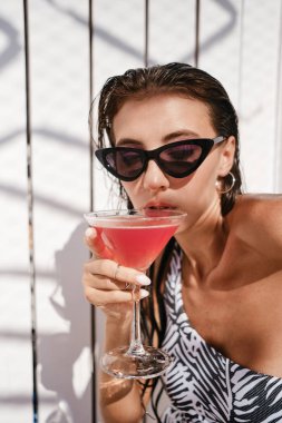 Tek parça mayo ve güneş gözlüğü çekici esmer kız Portresi düşünceli plajda ahşap arka plan üzerinde kokteyl içme