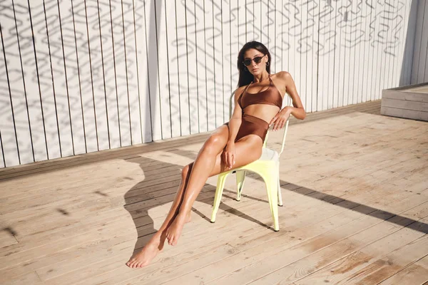 Vacker brunett flicka i baddräkt och solglasögon omtänksamt tittar i kameran sola på stol på stranden — Stockfoto